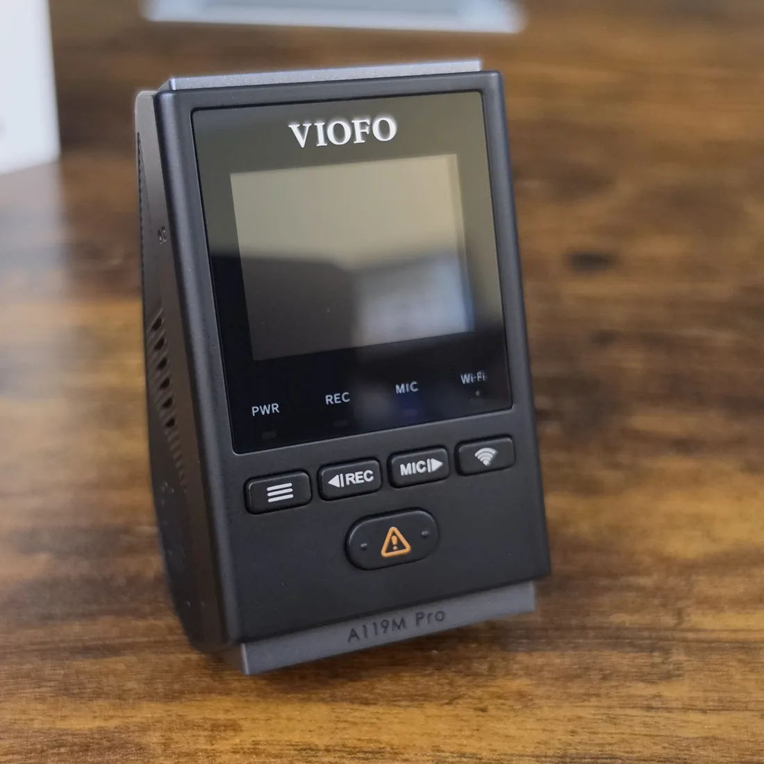The VIOFO A119M Pro mini dash cam standing on a wooden desk, its 1.5 inch screen, five buttons and the A119M Pro name on the mount rail facing the camera
